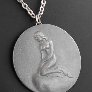 May include: A silver-toned pendant necklace featuring a large, round medallion. The medallion showcases a sculpted figure of a woman kneeling on a rock. The chain is made of interlocking oval links, and the background is black.