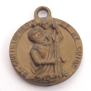 Op de afbeelding: Bronzen St. Christoffelmedaille met de tekst "ST. CHRISTOPHER BE MY GUIDE" langs de rand. De medaille toont St. Christoffel die een kind draagt. Het heeft een lus aan de bovenkant om op te hangen.