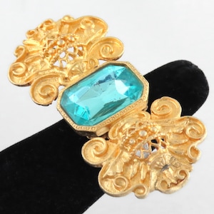 Może przedstawiać: Złota broszka z dużym, prostokątnym, turkusowym kamieniem osadzonym w środku. Broszka jest ozdobiona misternymi, wirującymi wzorami.