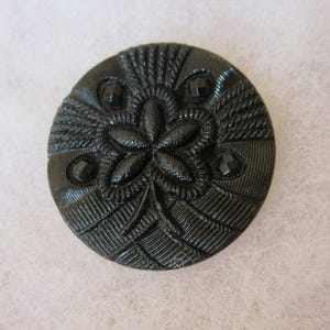 May include: A round black button featuring a central five-petalled floral motif surrounded by textured geometric patterns and four small faceted black accents. The surface displays intricate raised lines and a glossy finish throughout the design.