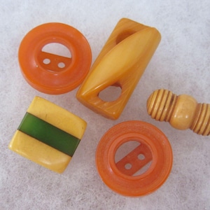 May include: A collection of five buttons including two bright orange circular buttons with two holes, one rectangular amber-coloured toggle button, one square yellow button with a green centre stripe, and one cylindrical cream-coloured ribbed button.