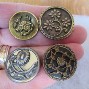 May include: Four circular metal buttons with intricate relief designs. The top left button features a floral motif in brass. The top right button has a dark finish with three star-like flowers. The bottom left is silver-toned with a sunflower, and the bottom right is a dark button with a metallic floral pattern.