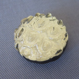 以下が含まれることがあります： テクスチャ加工された淡いベージュの表面と暗い縁を持つ、丸い装飾品。上部には渦巻き模様があり、オブジェクトは固い素材でできているようです。背景はグレーのテクスチャ加工された生地です。