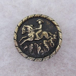 May include: A round button featuring a raised brass-toned relief of a person riding a horse on a black background. The circular border is decorated with a repeating leaf pattern in a brass tone, framing the central equestrian scene.