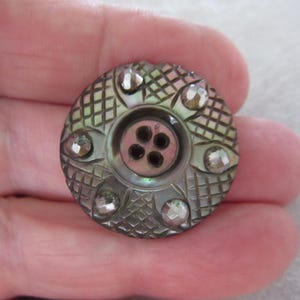 May include: A circular, vintage button with a mother-of-pearl finish. The button has a central design with four holes and is decorated with six clear, faceted rhinestones. The button has a detailed, textured pattern.