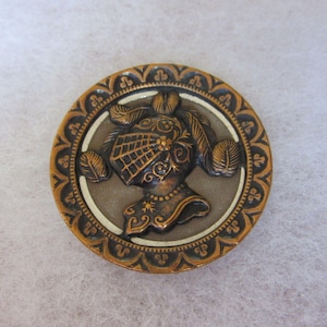 Peut inclure: Un bouton métallique rond présentant un relief représentant un casque de chevalier avec des détails de plumes. Le centre est d'un ton bronze sourd, entouré d'une bordure sombre et ornée avec des motifs floraux répétés en finition dorée.