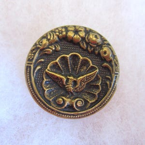 Peut inclure: Bouton rond en laiton antique avec un design détaillé. Le bouton présente un emblème central d'un oiseau aux ailes déployées, entouré d'un motif en forme d'éventail. Des détails floraux et des volutes ornent le bord du bouton.