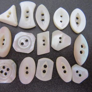 May include: A collection of fourteen white mother-of-pearl buttons in various geometric shapes, including triangles, ovals, rectangles, and squares. Each button features two or four central holes for attachment, showcasing a smooth, iridescent finish.
