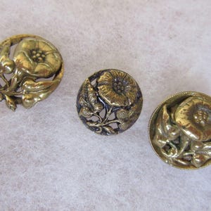 May include: Three circular brass buttons featuring a raised relief design of a flower with petals and leaves. The metal has a polished gold-toned finish with darkened recessed areas that highlight the intricate floral details and textures.