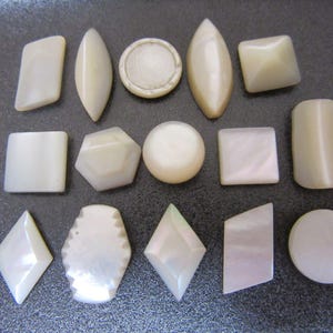 May include: A collection of fourteen off-white mother-of-pearl buttons in various geometric shapes, including circles, squares, diamonds, and elongated ovals. Each button features a smooth, iridescent surface with a subtle pearlescent sheen.
