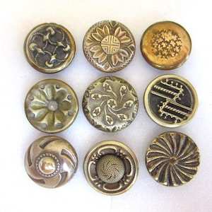 May include: A collection of nine antique brass buttons, each with a unique embossed design. The buttons feature floral, geometric, and abstract patterns, arranged in a 3x3 grid. The aged patina highlights their vintage charm, perfect for sewing and craft projects.
