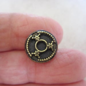 May include: A small, circular button with a dark grey centre and a decorative gold-coloured rim. The button features a gold-coloured floral design in the centre and around the edge. The button is likely made of metal.