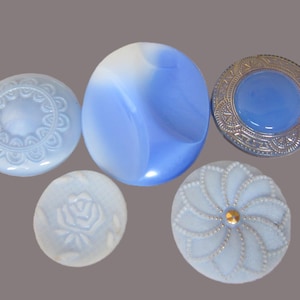May include: A collection of five round buttons in shades of blue and white. The set includes a light blue button with a scalloped edge, a large blue and white swirled button, a metallic-rimmed blue button, a small white rose-embossed button, and a light blue button with a beaded floral pattern and a small gold centre.
