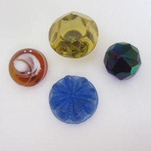 May include: Four decorative glass buttons in various colours. One is amber, faceted. Another is dark blue with a floral pattern. A third is dark, iridescent. The last button has a brown and white swirl design.