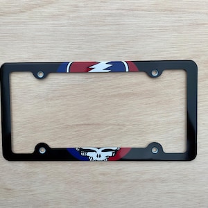 May include: Black license plate frame with a colourful design. The top and bottom feature a red, white, and blue pattern with a lightning bolt and a skull. The frame has four mounting holes.