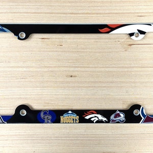 Op de afbeelding: Zwarte kentekenplaathouder met een witte rand en verschillende logo's van sportteams uit Denver, waaronder de Broncos, Nuggets, Rockies en Avalanche.