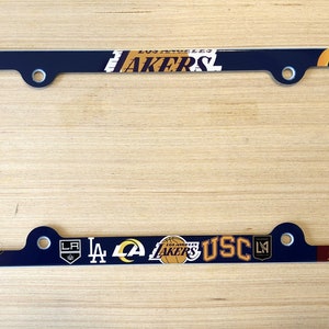 May include: A blue license plate frame with various Los Angeles sports team logos, including the Lakers, Rams, Dodgers, Kings, and USC Trojans. The frame is made of metal and has a glossy finish.