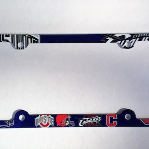 May include: Blue and red license plate frame with various sports team logos, including the Cleveland Cavaliers, Ohio State Buckeyes, and the Cleveland Indians. The frame has two holes on each side for mounting.
