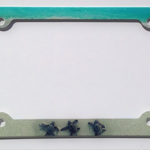 May include: A white license plate frame with a beach scene and three small sea turtles. The frame has a turquoise blue ocean wave at the top and a sandy beach at the bottom.