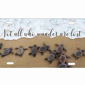 May include: A license plate with a beach scene featuring a wave and baby sea turtles walking on the sand. The text "Not all who wander are lost" is written in cursive on the plate.
