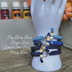 Puede incluir: Cuatro pulseras azules en diferentes tonos, incluyendo Icey Baby Blue, Royal Blue, Denim Steel Blue y Navy Blue, se exhiben en un soporte blanco con forma de mano. Cada pulsera presenta un anillo plateado y cuentas de madera. La imagen tambi&eacute;n incluye texto con los nombres de los colores de las pulseras.