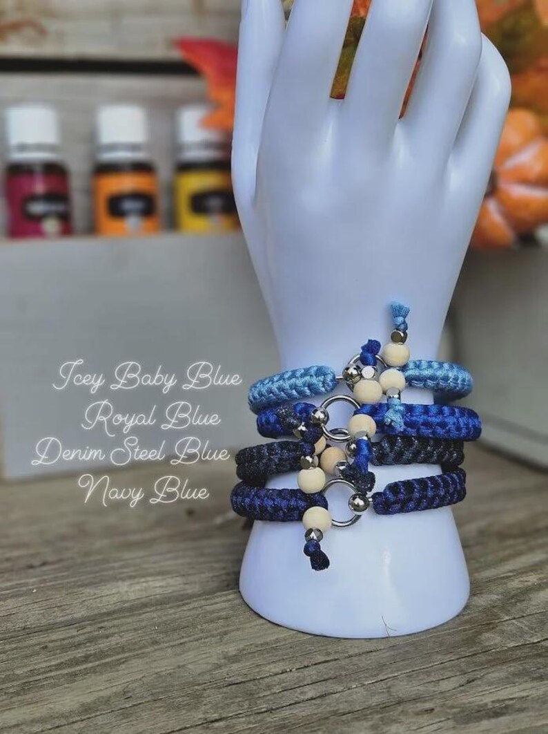 Puede incluir: Cuatro pulseras azules en diferentes tonos, incluyendo Icey Baby Blue, Royal Blue, Denim Steel Blue y Navy Blue, se exhiben en un soporte blanco con forma de mano. Cada pulsera presenta un anillo plateado y cuentas de madera. La imagen tambi&eacute;n incluye texto con los nombres de los colores de las pulseras.