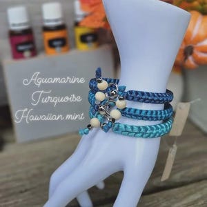 Puede incluir: Tres pulseras trenzadas en tonos azules, adornadas con cuentas de madera y detalles plateados, se exhiben en una mano de maniqu&iacute; blanca. Las pulseras est&aacute;n etiquetadas como "Aquamarine, Turquoise, Hawaiian mint."