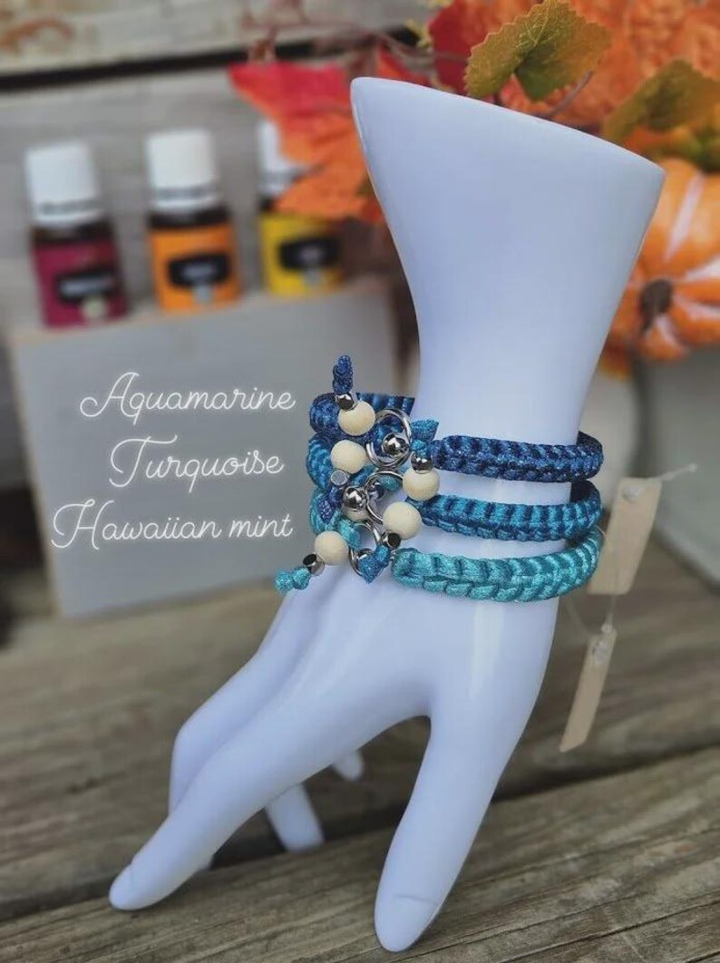 Puede incluir: Tres pulseras trenzadas en tonos azules, adornadas con cuentas de madera y detalles plateados, se exhiben en una mano de maniqu&iacute; blanca. Las pulseras est&aacute;n etiquetadas como "Aquamarine, Turquoise, Hawaiian mint."