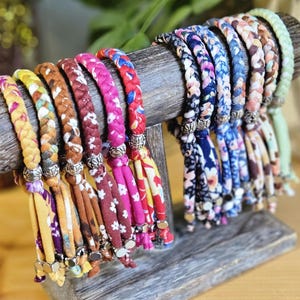 May include: A collection of braided fabric bracelets in various colors including yellow, brown, red, pink, and blue, featuring floral patterns. Each bracelet is accented with silver-toned metal beads and ends in loose fabric tassels with metal beads.