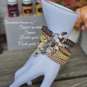 Puede incluir: Pulseras trenzadas en tonos marr&oacute;n chocolate, cobre y dorado, adornadas con cuentas de madera y detalles plateados, expuestas sobre una forma de mano blanca. El texto en la imagen dice: "Chocolate brown w/ Copper sparkle Copper Butter gold Nude gold."