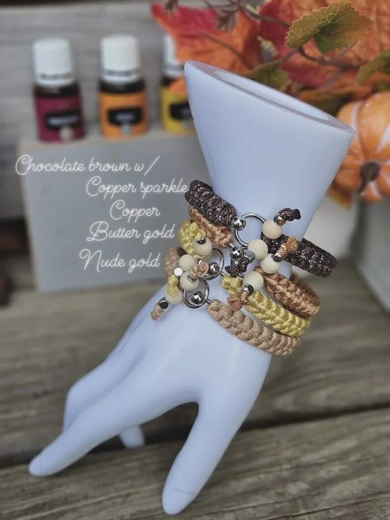 Puede incluir: Pulseras trenzadas en tonos marr&oacute;n chocolate, cobre y dorado, adornadas con cuentas de madera y detalles plateados, expuestas sobre una forma de mano blanca. El texto en la imagen dice: "Chocolate brown w/ Copper sparkle Copper Butter gold Nude gold."