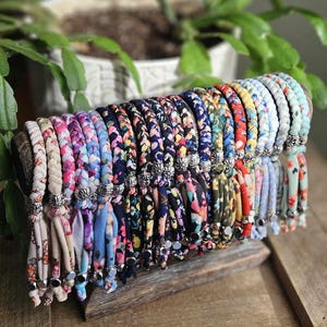 May include: A display of colorful fabric bracelets with floral patterns. The bracelets are arranged on a wooden stand, showcasing a variety of colors including pink, blue, and green. Each bracelet has a silver-colored metal accent and a tassel.