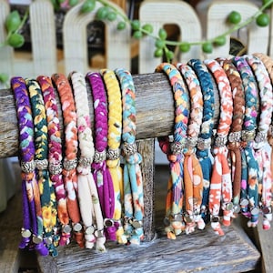 May include: A display of colorful braided fabric bracelets with floral and patterned designs. The bracelets are arranged on a wooden display stand, showcasing a variety of colors including purple, orange, blue, and yellow.