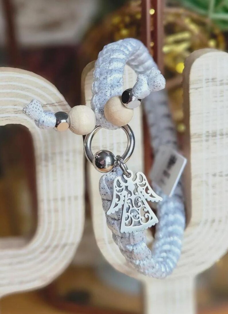 Puede incluir: Un llavero trenzado plateado y blanco con un dije decorativo de &aacute;ngel. El llavero presenta cuentas de madera y un anillo plateado. El dije de &aacute;ngel est&aacute; intrincadamente dise&ntilde;ado con detalles recortados.