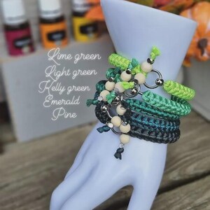 Puede incluir: Una colecci&oacute;n de pulseras de ganchillo verdes con cuentas de madera y anillos plateados, exhibidas en una mano blanca. Las pulseras son en tonos verde lima, verde claro, verde Kelly, esmeralda y pino. Tambi&eacute;n se ven botellas de aceites esenciales.