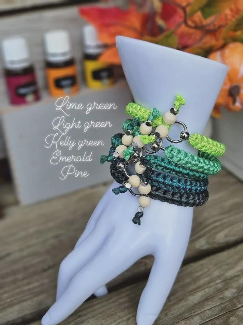 Puede incluir: Una colecci&oacute;n de pulseras de ganchillo verdes con cuentas de madera y anillos plateados, exhibidas en una mano blanca. Las pulseras son en tonos verde lima, verde claro, verde Kelly, esmeralda y pino. Tambi&eacute;n se ven botellas de aceites esenciales.