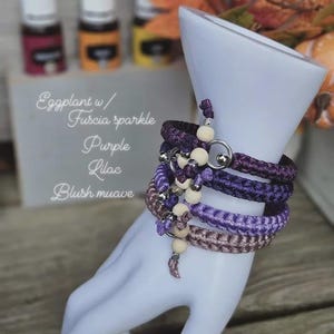 Puede incluir: Un conjunto de pulseras trenzadas en tonos morados y malva, adornadas con cuentas de madera y detalles met&aacute;licos plateados. Las pulseras se exhiben en una forma de mano blanca. Se ven botellas de aceites esenciales.