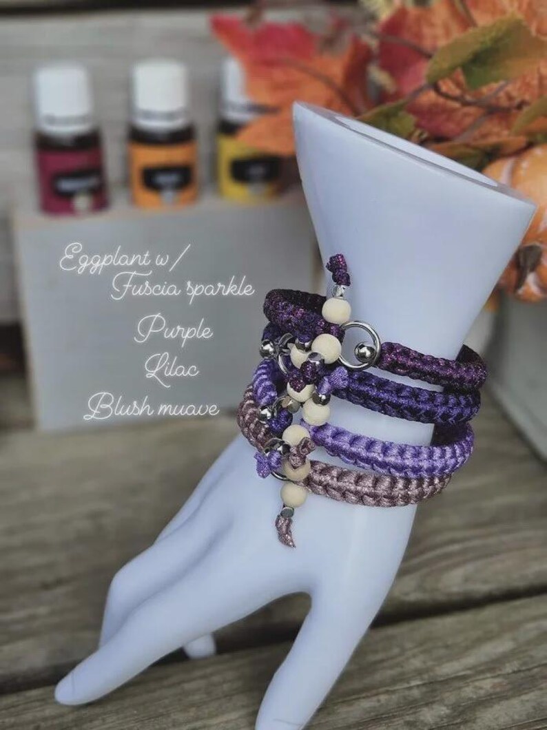 Puede incluir: Un conjunto de pulseras trenzadas en tonos morados y malva, adornadas con cuentas de madera y detalles met&aacute;licos plateados. Las pulseras se exhiben en una forma de mano blanca. Se ven botellas de aceites esenciales.