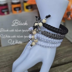 Puede incluir: Exhibici&oacute;n de pulseras trenzadas en negro, blanco y brillo plateado, adornadas con cuentas de madera y detalles plateados. Las pulseras se muestran en una mano de maniqu&iacute; blanca. El texto en la imagen dice "Black", "Black with silver sparkle", "White with silver sparkle" y "White".
