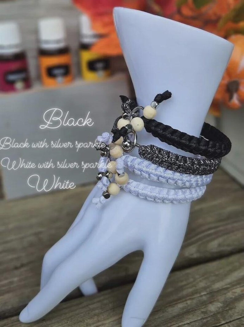 Puede incluir: Exhibici&oacute;n de pulseras trenzadas en negro, blanco y brillo plateado, adornadas con cuentas de madera y detalles plateados. Las pulseras se muestran en una mano de maniqu&iacute; blanca. El texto en la imagen dice "Black", "Black with silver sparkle", "White with silver sparkle" y "White".