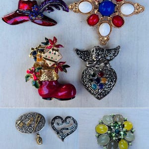 May include: Collection of vintage brooches in various shapes and designs. Includes a red hat with purple feather, a starburst with red, white, and blue stones, a red boot with gifts, a heart-shaped angel, and an elephant.