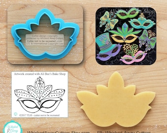 May include: A blue plastic cookie cutter shaped like a Mardi Gras mask. The cutter is on a wooden surface. The image also shows a decorated cookie in the shape of a Mardi Gras mask.