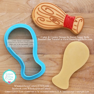 May include: A blue plastic cookie cutter shaped like a baseball bat. The cutter is on a wooden surface next to a decorated cookie in the shape of a baseball bat. The cookie is brown with red and white details. The text "Cutter design copyright protected by Whisked Away Cutters Cutter not to be recreated under U.S. & International Copyright Law" is visible on the image.
