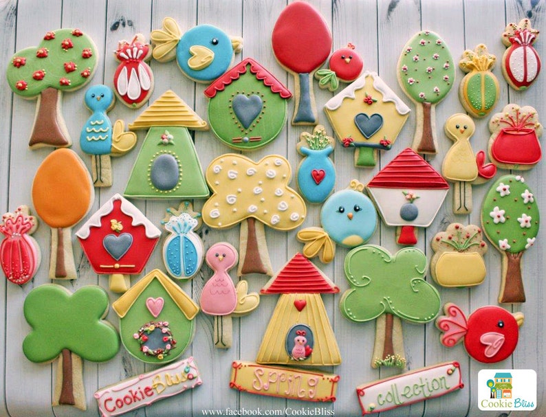 May include: A collection of decorated sugar cookies featuring birdhouses, trees, birds, and other spring-themed designs. The cookies are decorated with colorful icing and sprinkles. The text "Cookie Bliss Collection" is written on a banner at the bottom of the image.