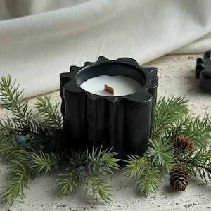 Peut inclure: Une bougie noire dans un support décoratif avec une bougie blanche et une mèche en bois. La bougie est entourée de branches de conifères, de petites pommes de pin et de baies bleues. Le bougeoir a un design texturé unique.