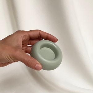 Peut inclure: Un petit porte-bougie en céramique de couleur vert clair, de forme ronde avec une dépression circulaire peu profonde au centre.