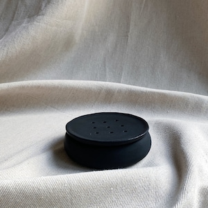 May include: A black ceramic soap dish with a round shape and multiple drainage holes. The dish is sitting on a white fabric surface.