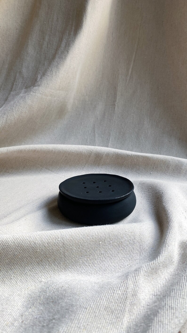 May include: A black ceramic soap dish with a round shape and multiple drainage holes. The dish is sitting on a white fabric surface.