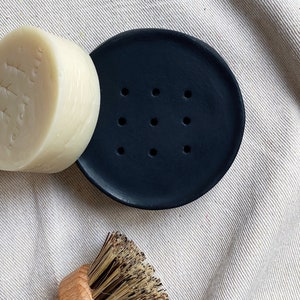 May include: A black ceramic soap dish with drainage holes, a white bar of soap, and a wooden brush with natural bristles.