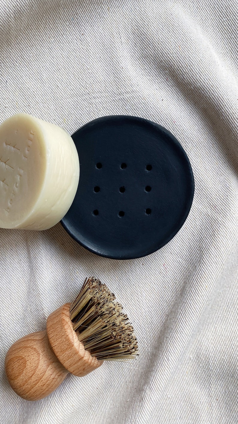 May include: A black ceramic soap dish with drainage holes, a white bar of soap, and a wooden brush with natural bristles.